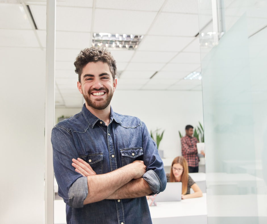A young man with curly hair and a beard, smiling confidently with arms crossed, standing in a bright modern office with blurred colleagues working in the background, illustrating a professional using tools like HubSpot's Prospecting Agent.
