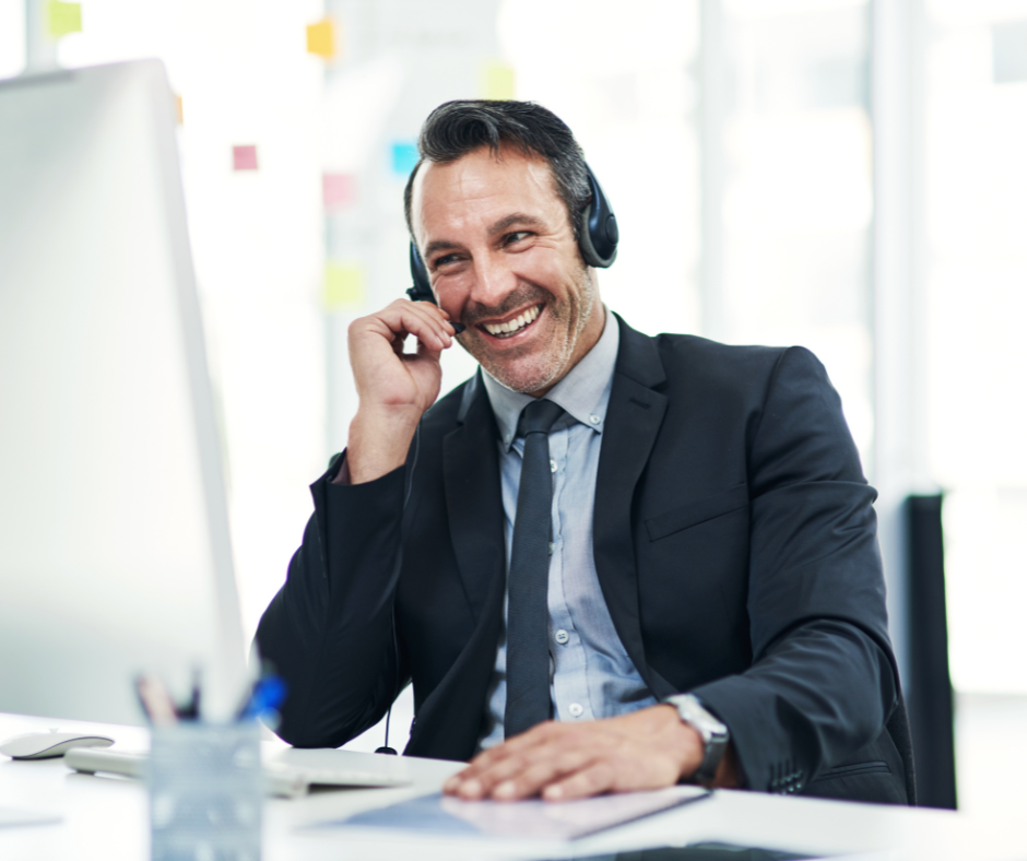 A middle-aged man in a business suit and tie, smiling while wearing a headset and speaking on a call at his desk with a computer monitor, representing successful lead engagement with tools like HubSpot's Prospecting Agent in a bright office setting.
