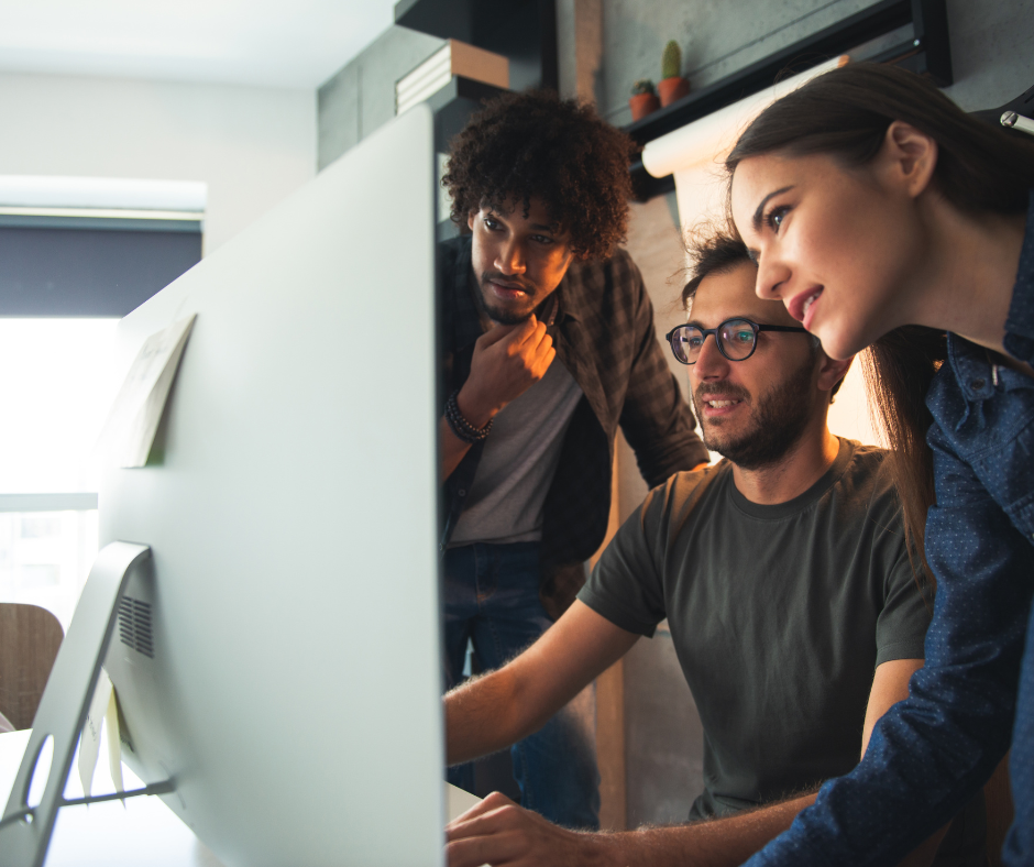 A diverse team of three young professionals—two men and one woman—gathered around a large computer monitor in a modern office, intently discussing and working on the screen, representing collaborative use of sales tools like HubSpot's Prospecting Agent.